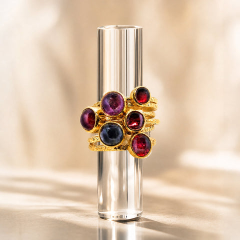 A gold ring with multiple round gemstones in deep red, purple, and dark blue, displayed on a clear cylindrical stand against a warm beige background.