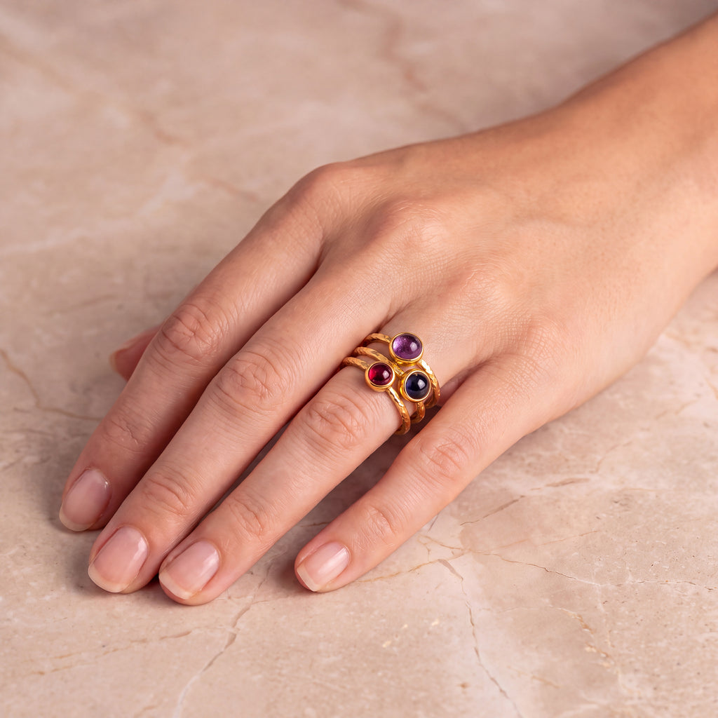 A gold ring with three small round gemstones in purple, red, and black, worn on a person's hand resting on a beige marble surface to show approximate scale