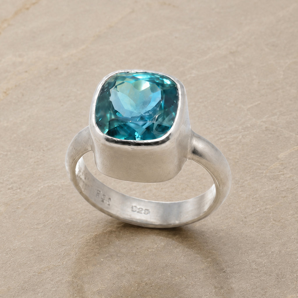 A sterling silver ring featuring a cushion-cut blue topaz gemstone, displayed on a neutral beige stone surface.
