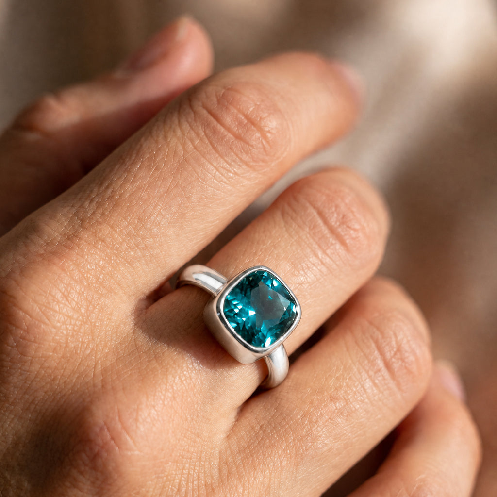A silver ring with a square-cut, cushion-cut turquoise gemstone worn on a person's finger, shown in a close-up shot with warm, natural lighting.