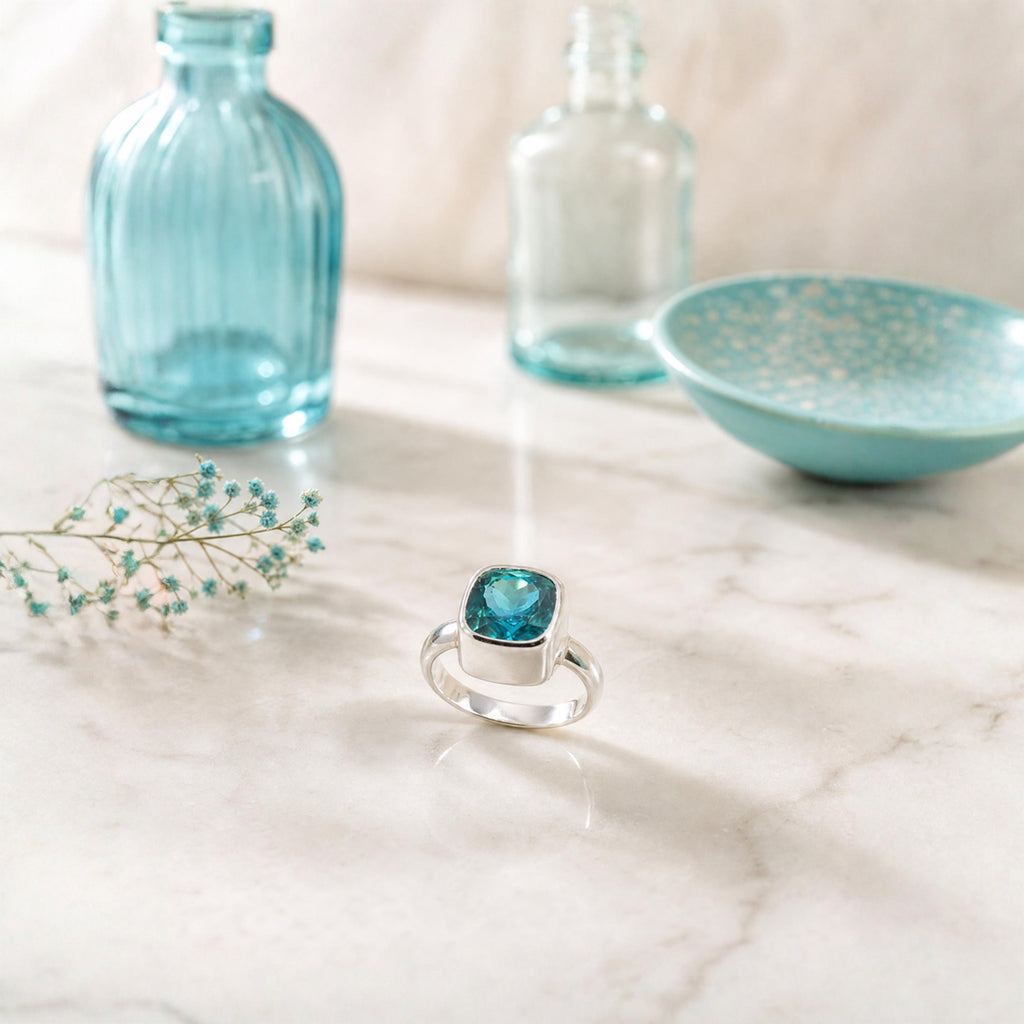 A silver ring with a rectangular bezel-set turquoise gemstone, displayed on a white marble surface with decorative blue glass bottles and a bowl in the background.