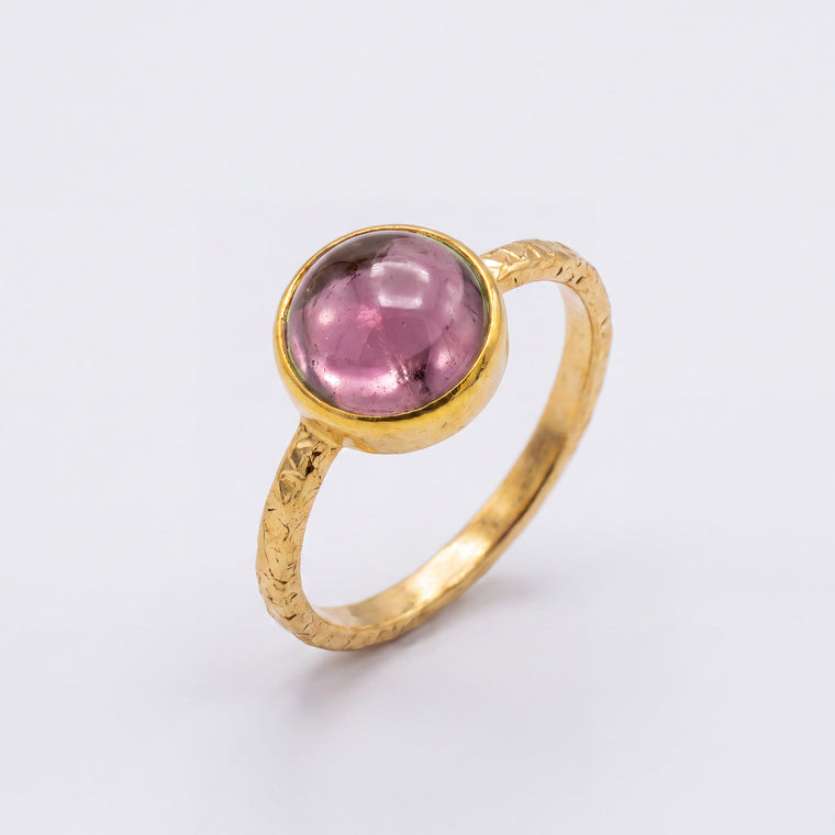 A gold ring featuring a round, faceted pink gemstone in a bezel setting, shown on a clean white background.