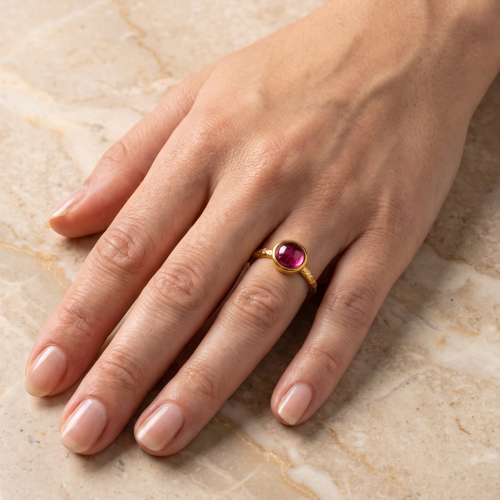 A gold ring with a round pink gemstone worn on a person's ring finger, shown against a light beige marble background, to show approximate scale.