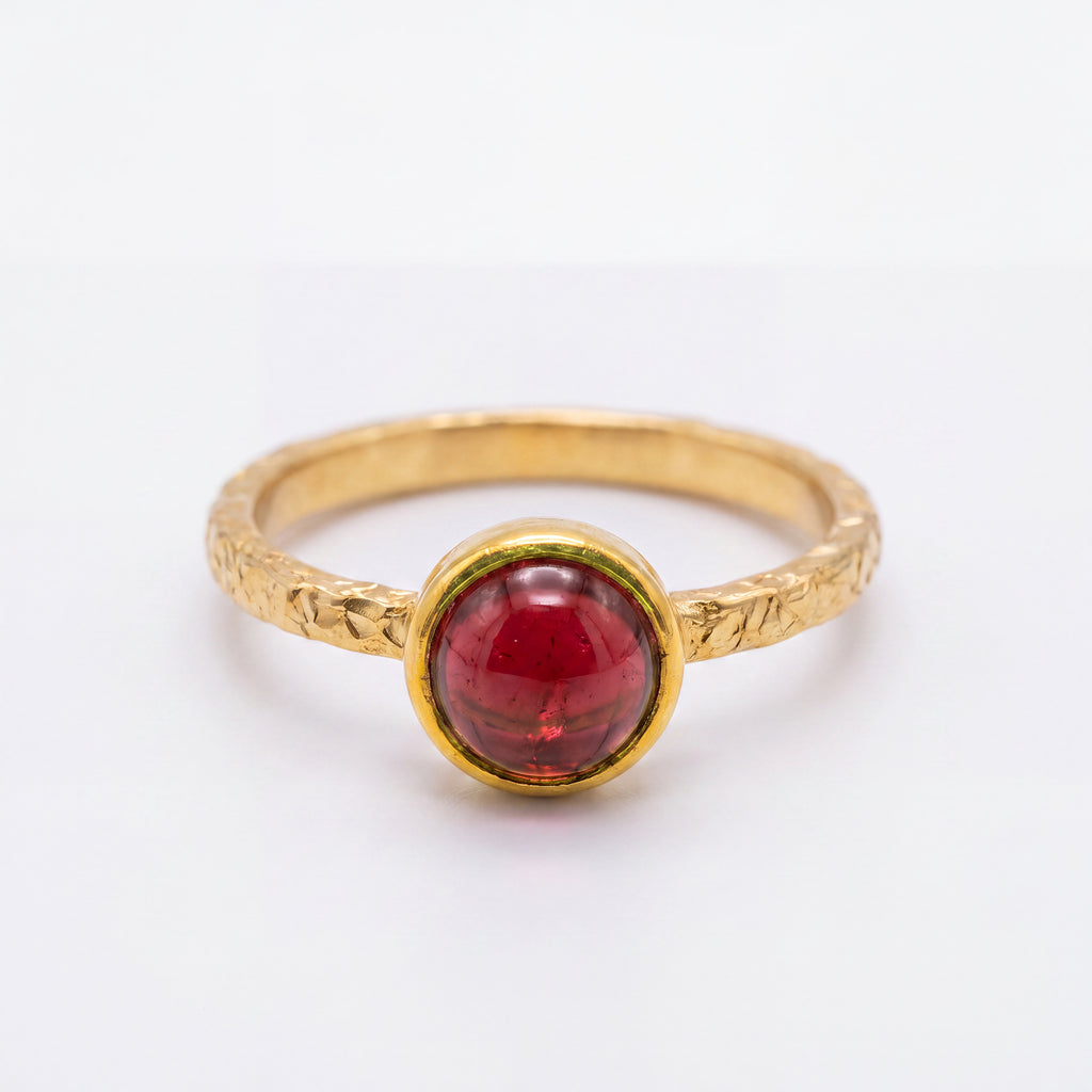 A gold ring with a textured band and a round, polished red gemstone in a bezel setting, shown on a plain white background.
