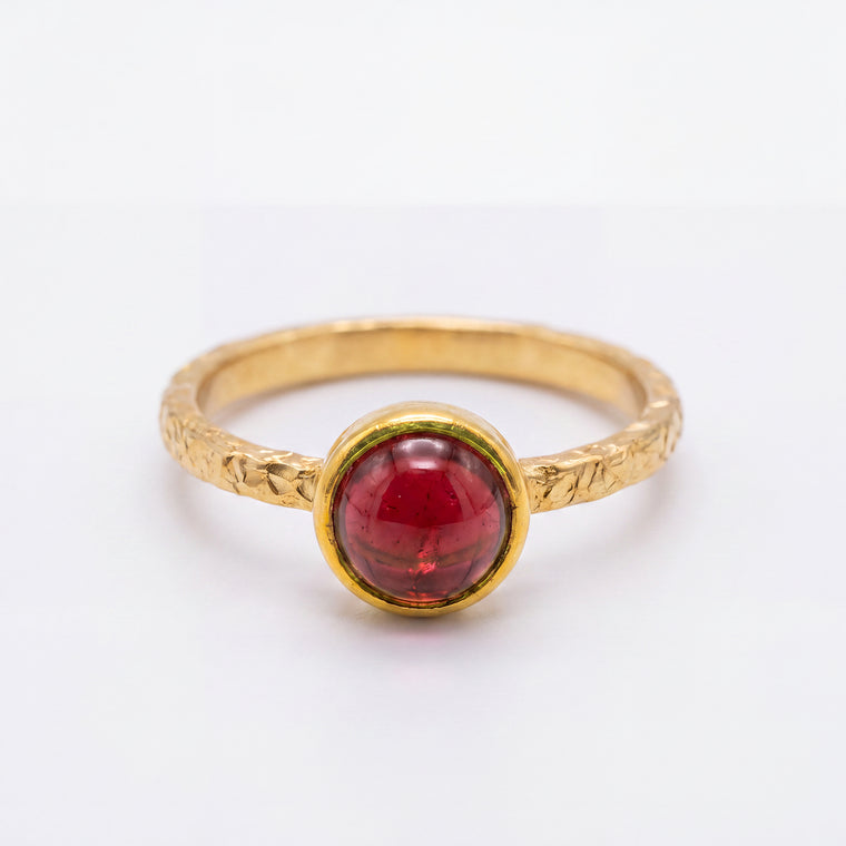 A gold ring with a textured band and a round, polished red gemstone in a bezel setting, shown on a plain white background.