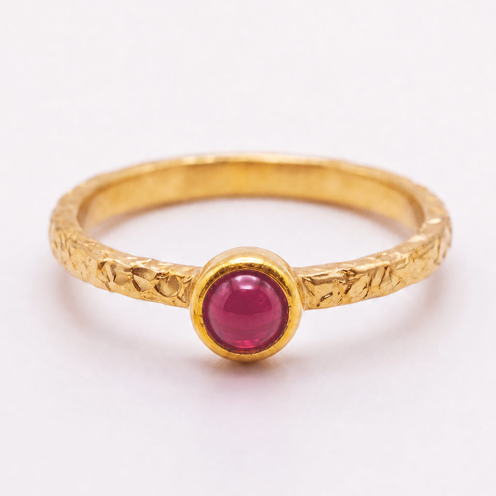A textured yellow gold ring featuring a round cabochon pink gemstone in a bezel setting, shown on a clean white background.