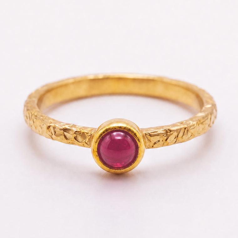 A textured yellow gold ring featuring a round cabochon pink gemstone in a bezel setting, shown on a clean white background.